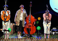 Szene aus dem Stück 'Hast du schon gehört?' des Theaters Marabu aus Bonn - Foto: Ursula Kaufmann Drei Schauspieler mit Instrumenten auf einer Bühne mit großen Blumen