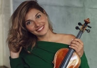 Eine junge Frau mit braunen Haaren und einer Geige blickt lächelnd in die Kamera. 