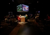 Eine dunkle Bühne mit einem beleuchteten großen Bildschirm und Menschen, die an Tischen vor Rechnern sitzen.