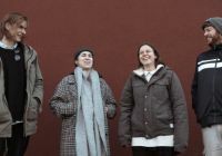 Vier Personen, zwei Männern und zwei Frauen, stehen vor einer roten Wand und lachen. 